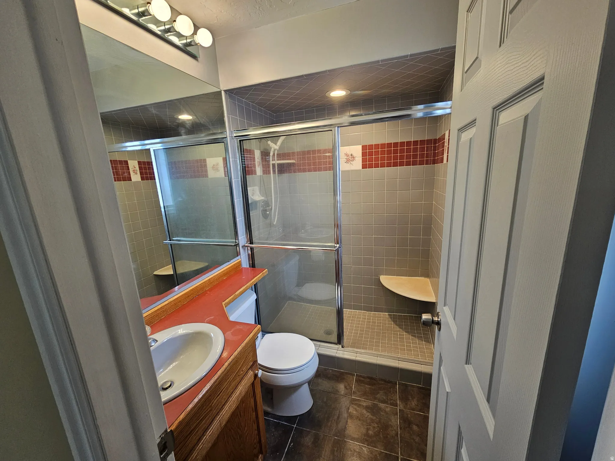 Bathroom featuring a wood-finish vanity with an integrated sink and a red countertop, a full-wall mirror, and a shower enclosure with a built-in bench