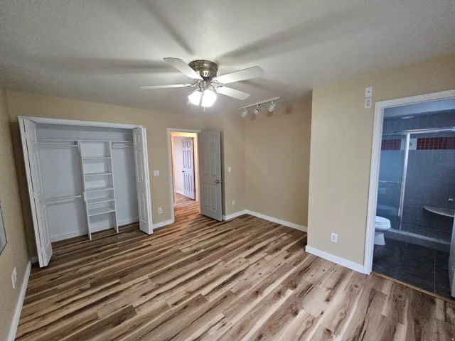 Spacious room featuring wood-finish flooring, a ceiling fan with integrated lighting, track lighting fixtures, and a built-in closet system with shelving and hanging rods