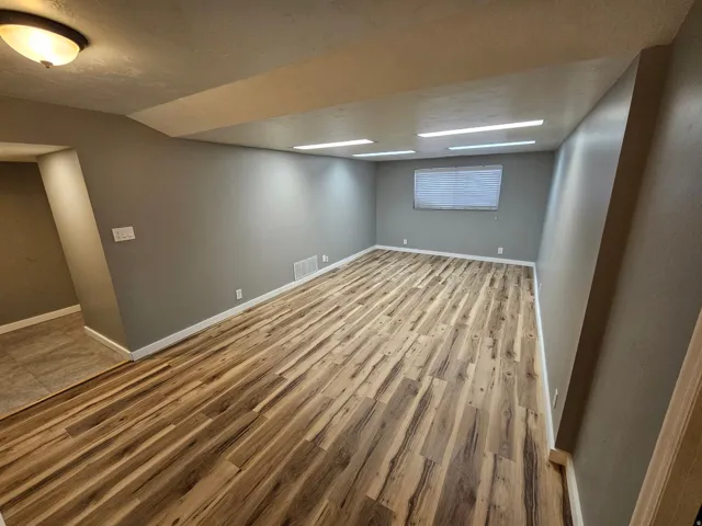 Spacious room featuring wood-finish flooring, neutral gray walls, a window with blinds, and white trim