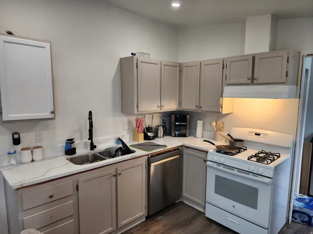 L-shaped kitchen featuring white quartz-look countertops, a stainless steel double-basin sink with a matte black faucet, and a full suite of white appliances