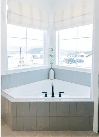Corner soaking tub featuring matte black fixtures, light grey vertical tile surround, and two expansive windows with striped Roman shades