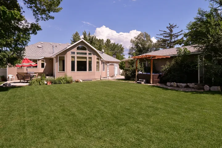 Expansive backyard featuring a manicured lawn, a large patio area, a dedicated hot tub area with pergola, mature perimeter trees, and a stucco exterior with an arched window design