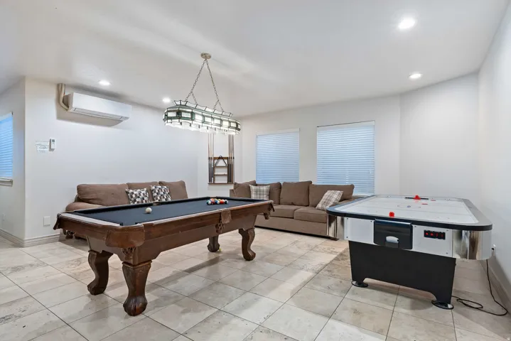 Spacious recreation area featuring a detailed billiards table under a distinctive chandelier, light-toned tile flooring, recessed lighting, and multiple windows with blinds