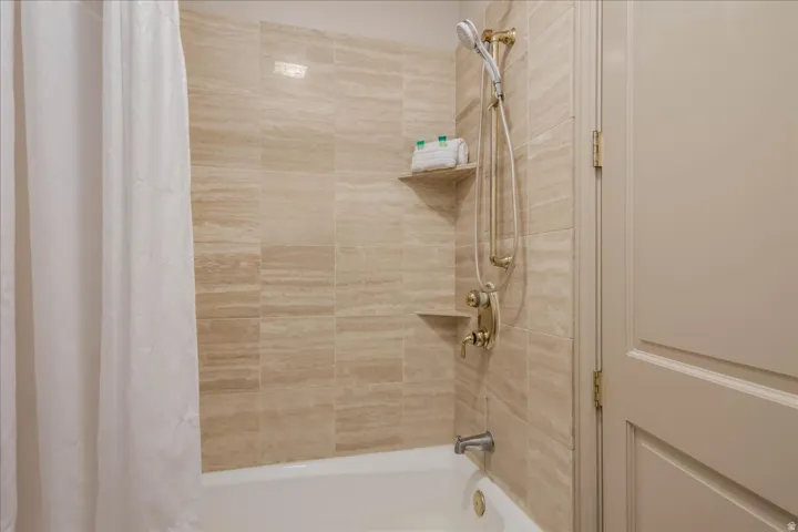 Bathtub-shower combination featuring a fixed showerhead with an additional handheld sprayer, integrated shelving, and beige-toned tile surround