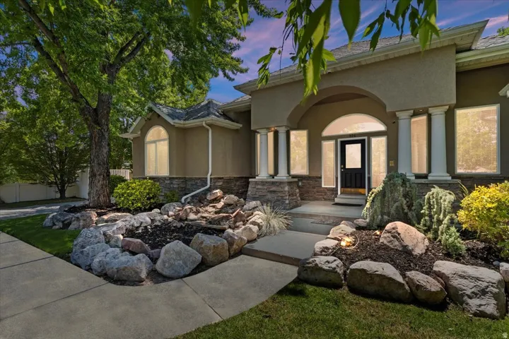 Stucco exterior featuring stacked stone accents, an arched entryway with decorative columns, mature tree landscaping, a concrete walkway, and exterior lighting