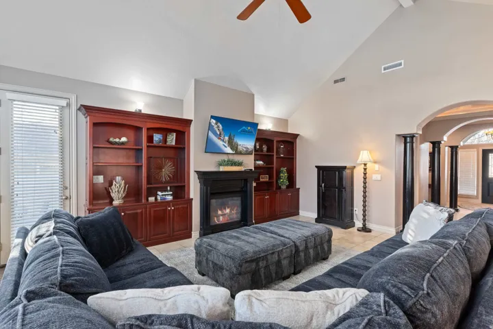Vaulted ceiling living area featuring a built-in fireplace with a hearth, custom built-in shelving, wood-finish ceiling fan, and architectural archways