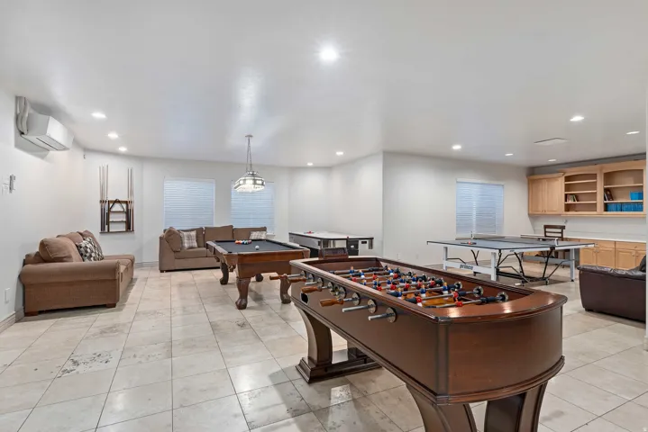 Spacious recreation room featuring light-toned tile flooring, recessed lighting, and a built-in cabinetry unit with open shelving