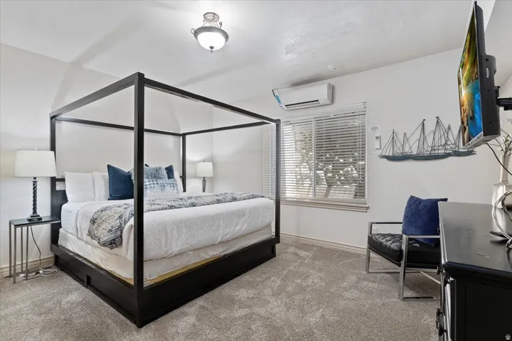 Spacious bedroom featuring neutral carpeting, white walls, a mounted air conditioning unit, a window with blinds, and a semi-flush mount ceiling light fixture