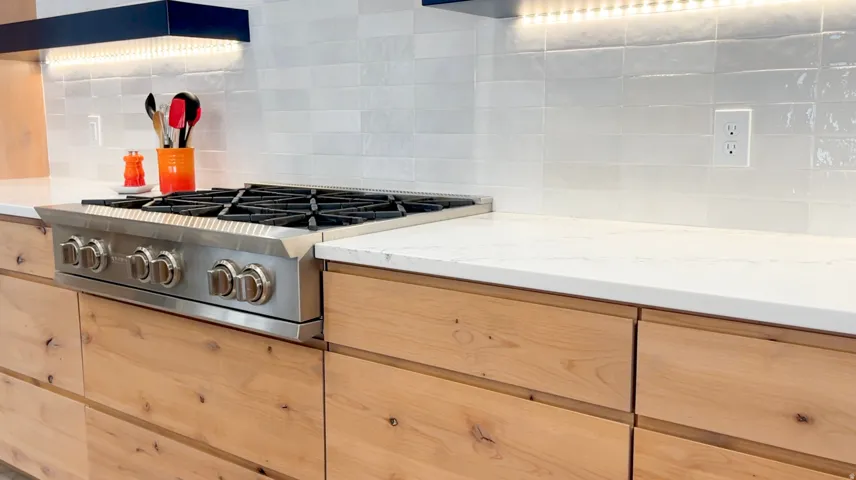 Stainless steel range with cast iron grates, light wood-finish cabinetry, white solid surface countertops, white subway tile backsplash, and under-cabinet lighting