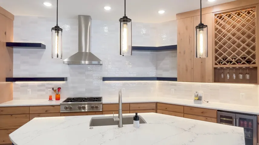 Modern kitchen featuring a large central island with an undermount sink, stainless steel appliances, a built-in wine rack with glass storage, light wood cabinetry, and white subway tile backsplash with under-cabinet lighting