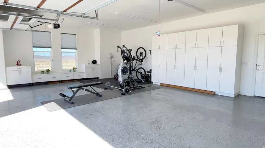 Spacious garage featuring extensive built-in white cabinetry, two large windows with horizontal blinds, a durable epoxy-coated concrete floor, and a white entry door