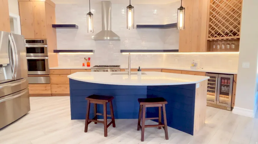 Kitchen featuring a curved island with a blue base, light wood-finish cabinetry, stainless steel appliances, a tile backsplash, and a built-in wine rack