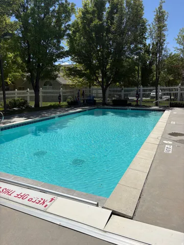 Rectangular swimming pool with light blue water surrounded by concrete decking and a white picket fence