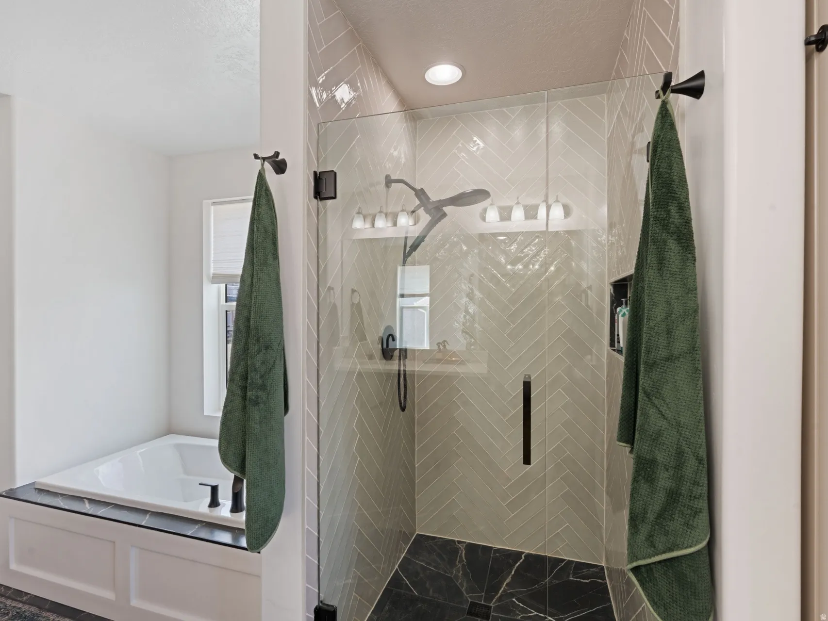 Primary bathroom with Glass-enclosed shower with herringbone tile surround and dark stone base, complemented by a built-in soaking tub with dark stone decking