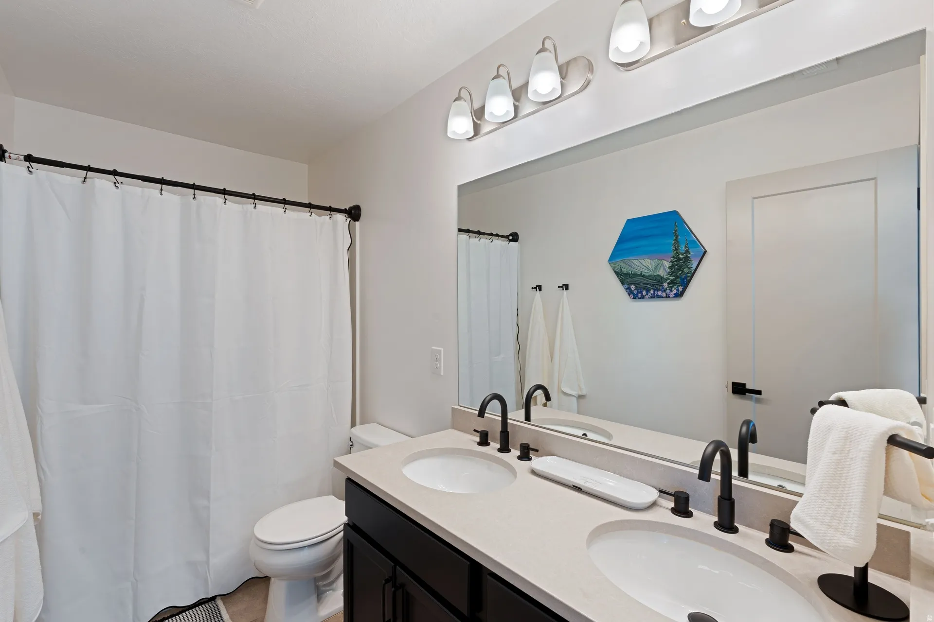Bathroom featuring a dual vanity with light-colored countertops, matte black fixtures, a large wall mirror, and a shower with a white curtain