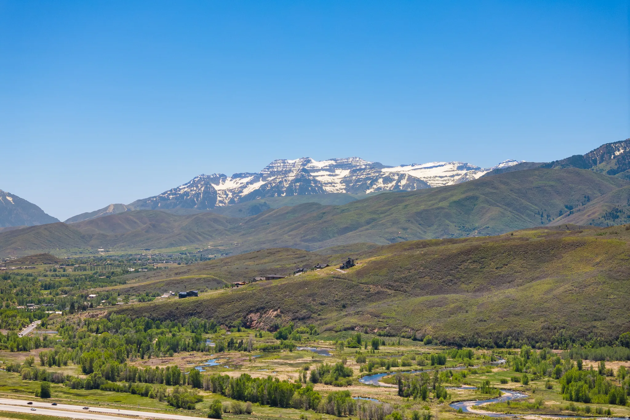 Expansive natural landscape featuring snow-capped mountains, rolling foothills, a winding river, and distant green valleys