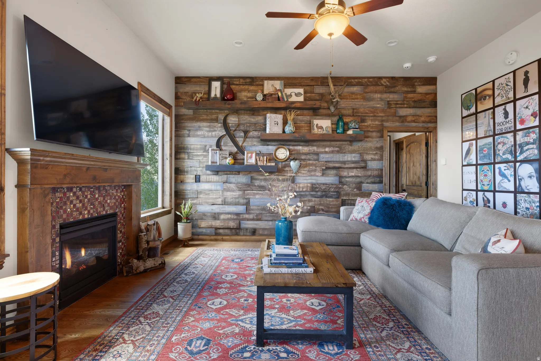Living area featuring a floor-to-ceiling wood plank accent wall, a fireplace with a wood mantle and mosaic tile surround, wood-finish flooring, a ceiling fan, and built-in shelving