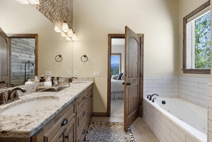 Bathroom featuring a dual vanity with granite-style countertops and undermount sinks, a built-in tub with tile surround, a large window, and a wooden interior door