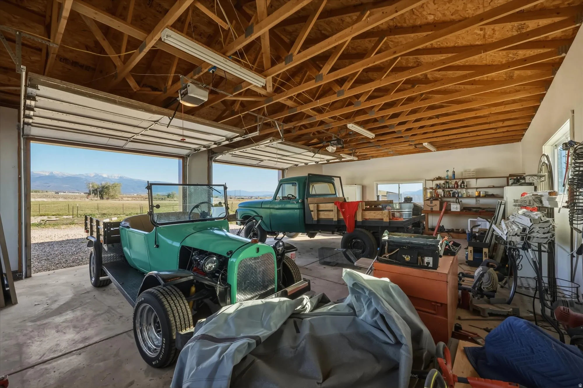 Spacious garage featuring exposed wood trusses, multiple overhead fluorescent light fixtures, two large roll-up garage doors, and concrete flooring
