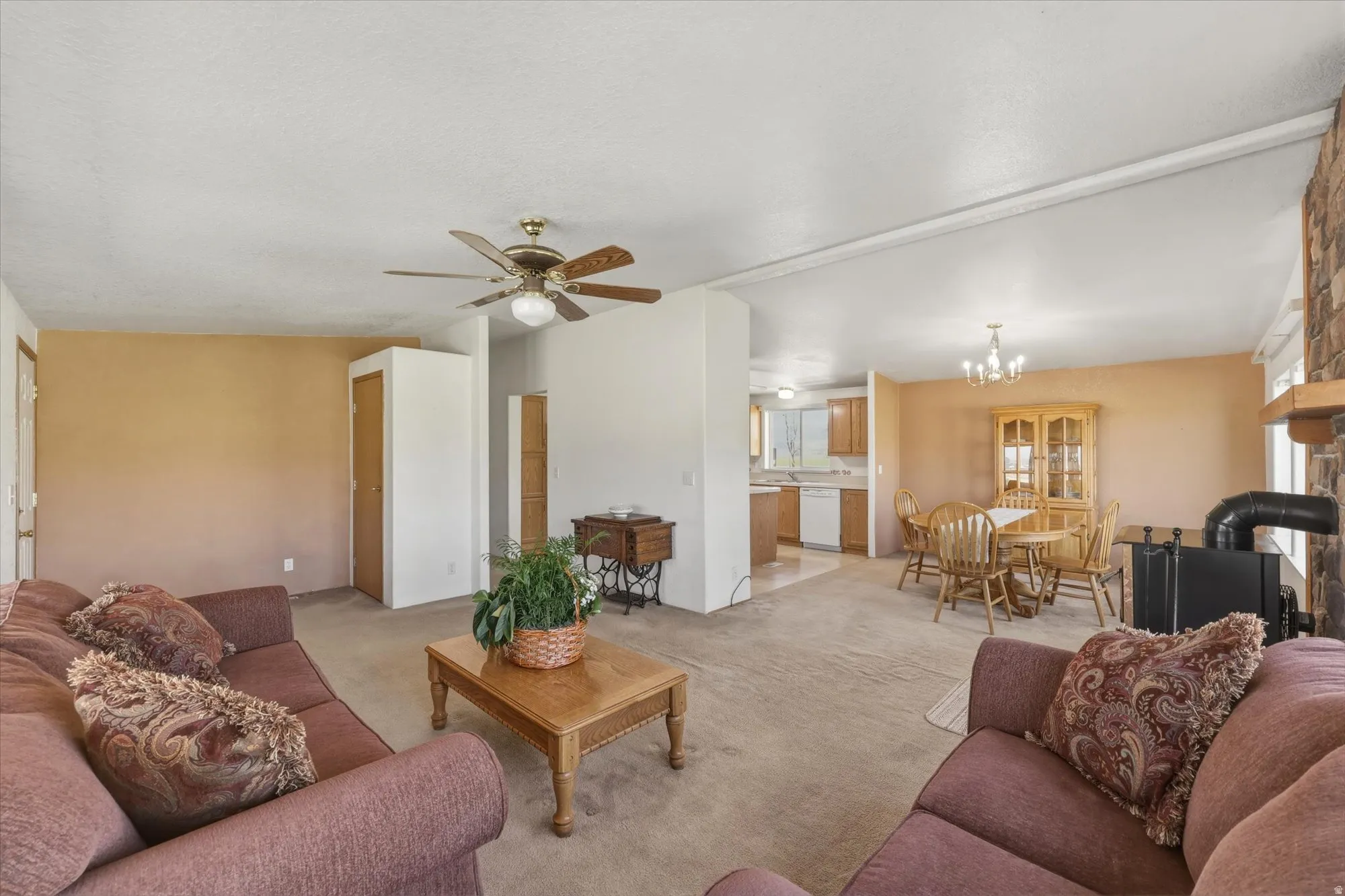 Open-concept living space featuring a ceiling fan, a kitchen with wood-finish cabinetry, a dining area with a chandelier, a stone-clad hearth, and a freestanding stove