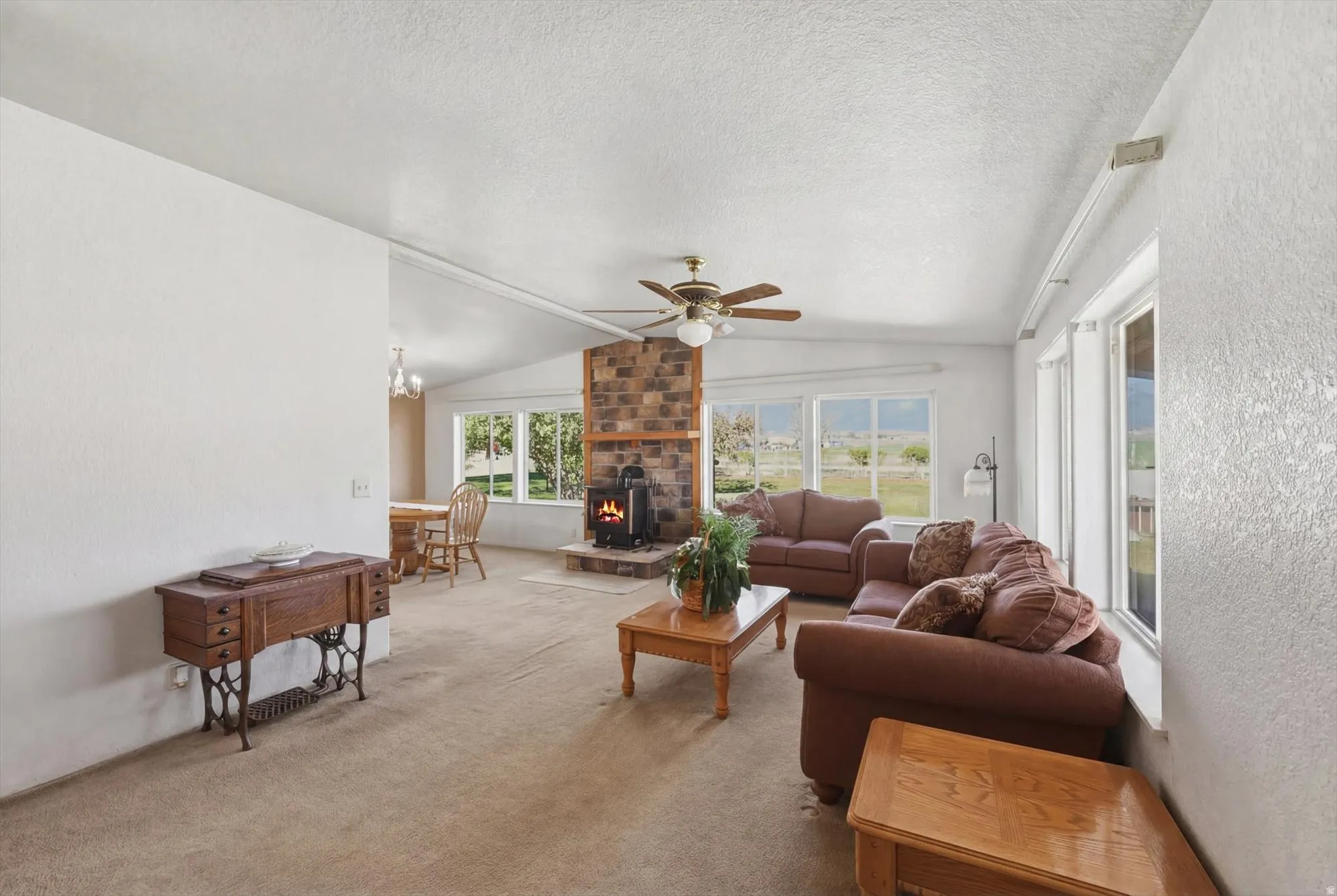 Living area featuring a stone-clad fireplace with a wood-burning stove, multiple windows, a ceiling fan, and sloped ceilings