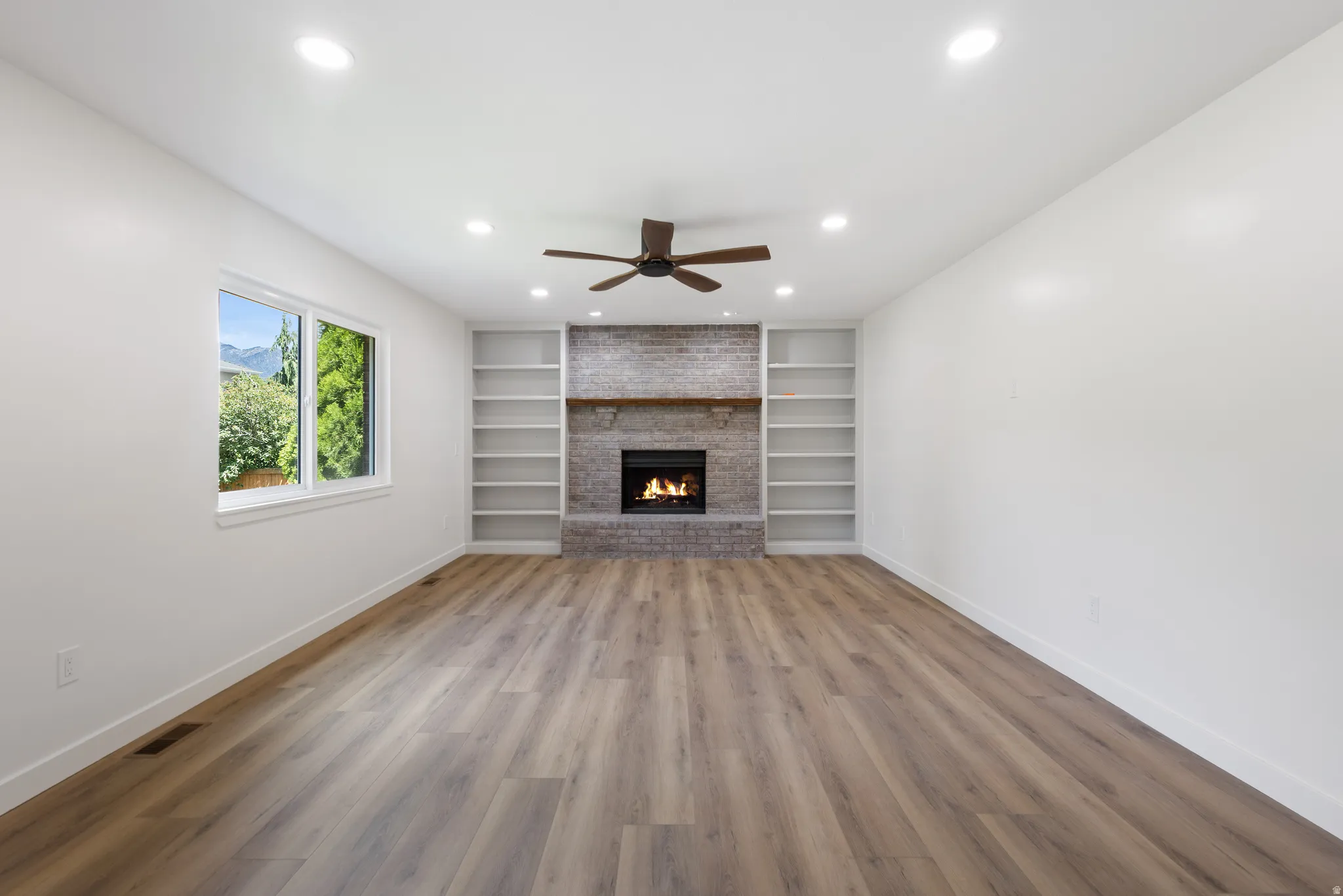 Spacious room featuring wood-finish flooring, a brick fireplace with a wood mantle, built-in shelving, a ceiling fan, and recessed lighting
