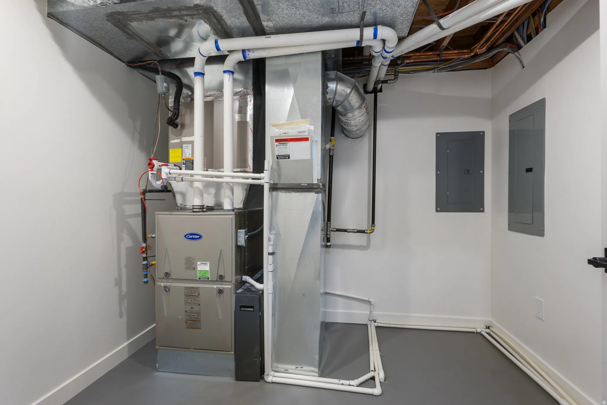 Utility area featuring a Carrier furnace system, exposed overhead ductwork, two electrical panels, and a painted concrete floor