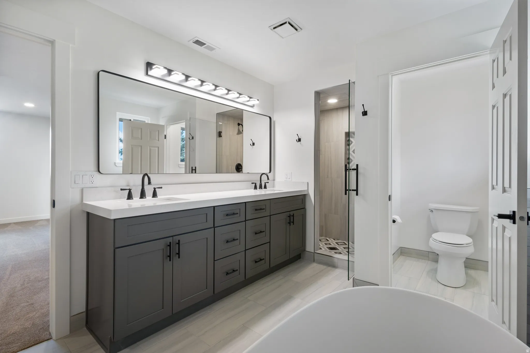 Modern bathroom featuring a dual vanity with a white countertop and dark cabinetry, a frameless glass shower enclosure, a freestanding tub, and light-toned tile flooring