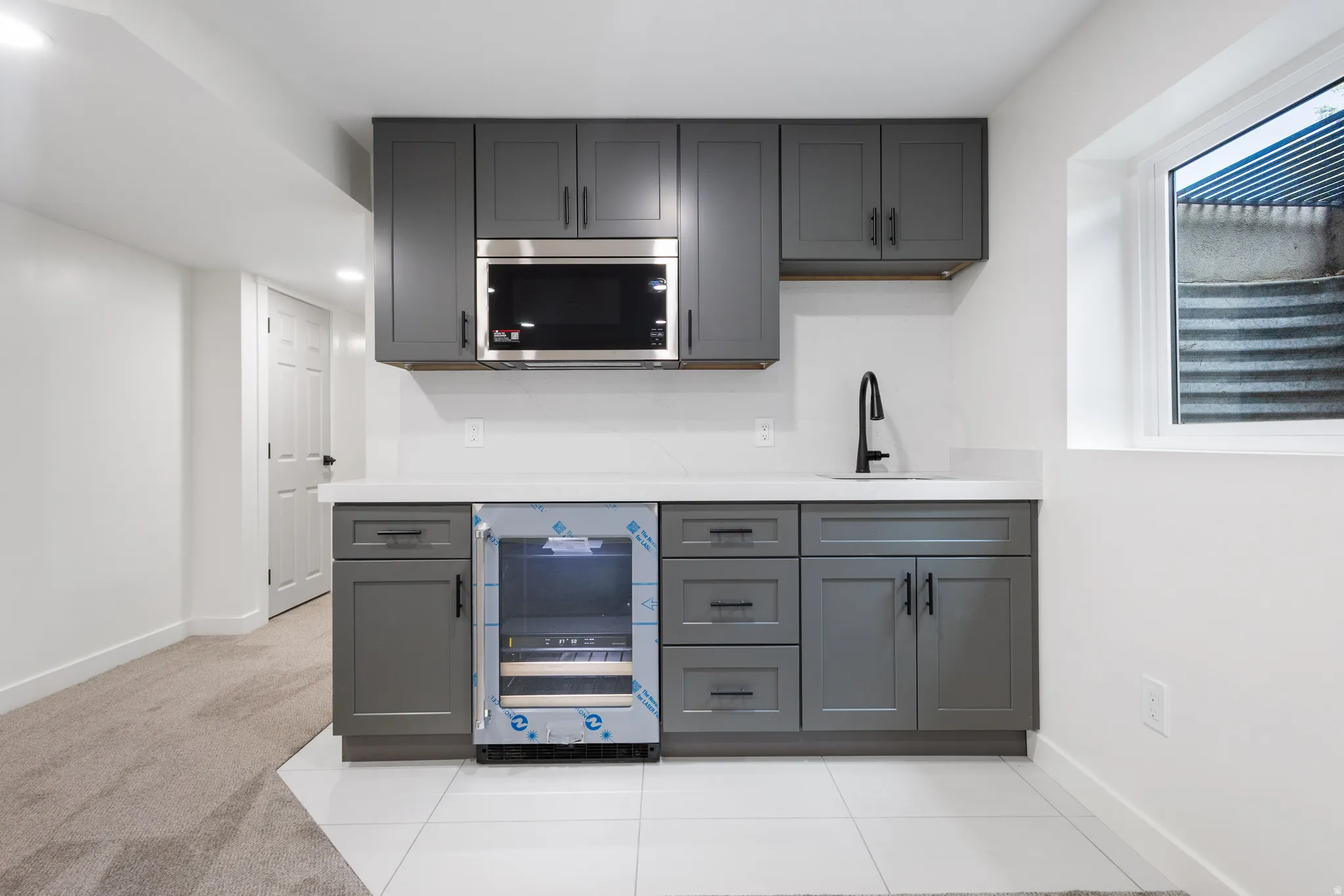 Modern wet bar featuring dark gray shaker cabinetry, a white countertop, stainless steel microwave, black faucet, and under-counter beverage refrigerator