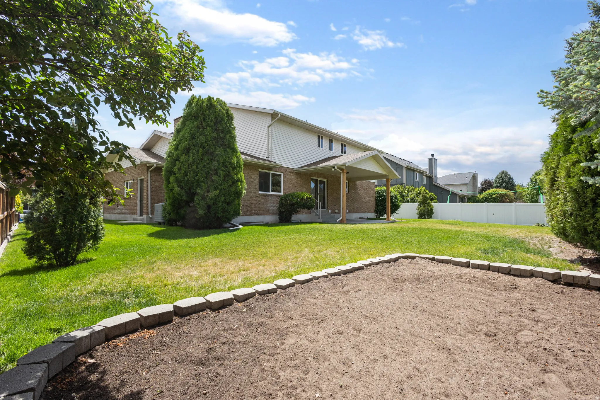 Expansive backyard featuring a large green lawn, a covered patio, brick and siding exterior, established perimeter trees, and a defined garden bed with stone edging