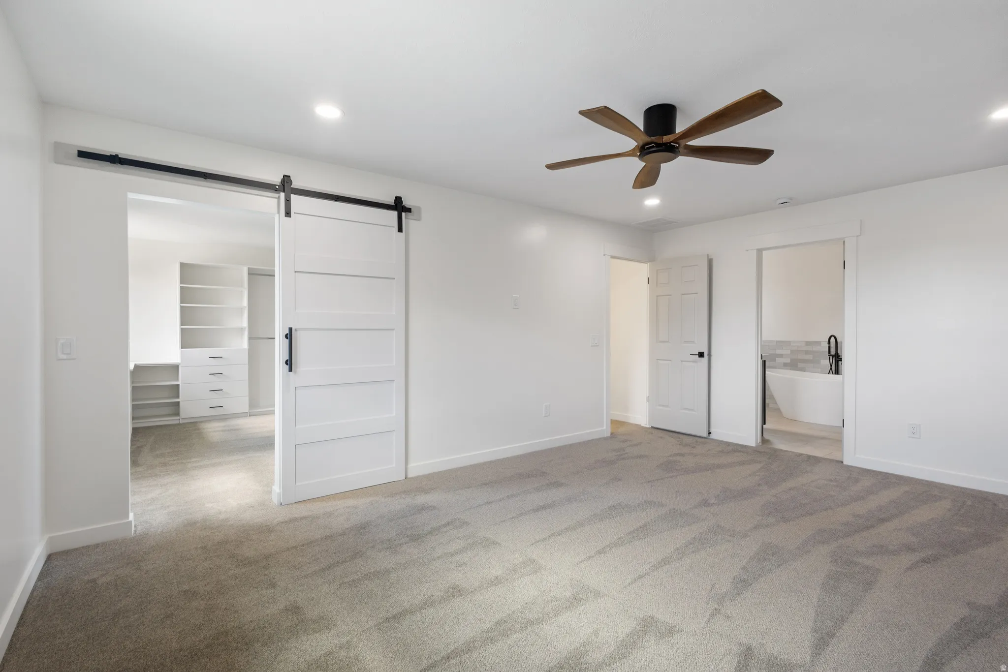 Spacious bedroom featuring plush gray carpeting, a contemporary wood-finish ceiling fan, recessed lighting, and a modern barn door opening to a walk-in closet with built-in shelving and drawers