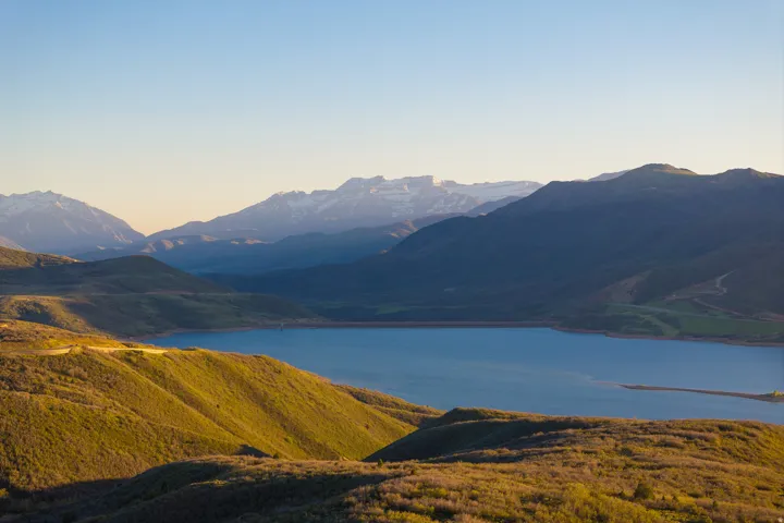 Expansive mountain landscape featuring a large body of water, rolling hills with varied foliage, and distant snow-capped peaks under a clear sky