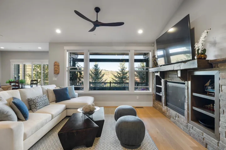 Spacious living area featuring expansive windows, a modern ceiling fan, wood-finish flooring, and a stone-clad fireplace with built-in shelving