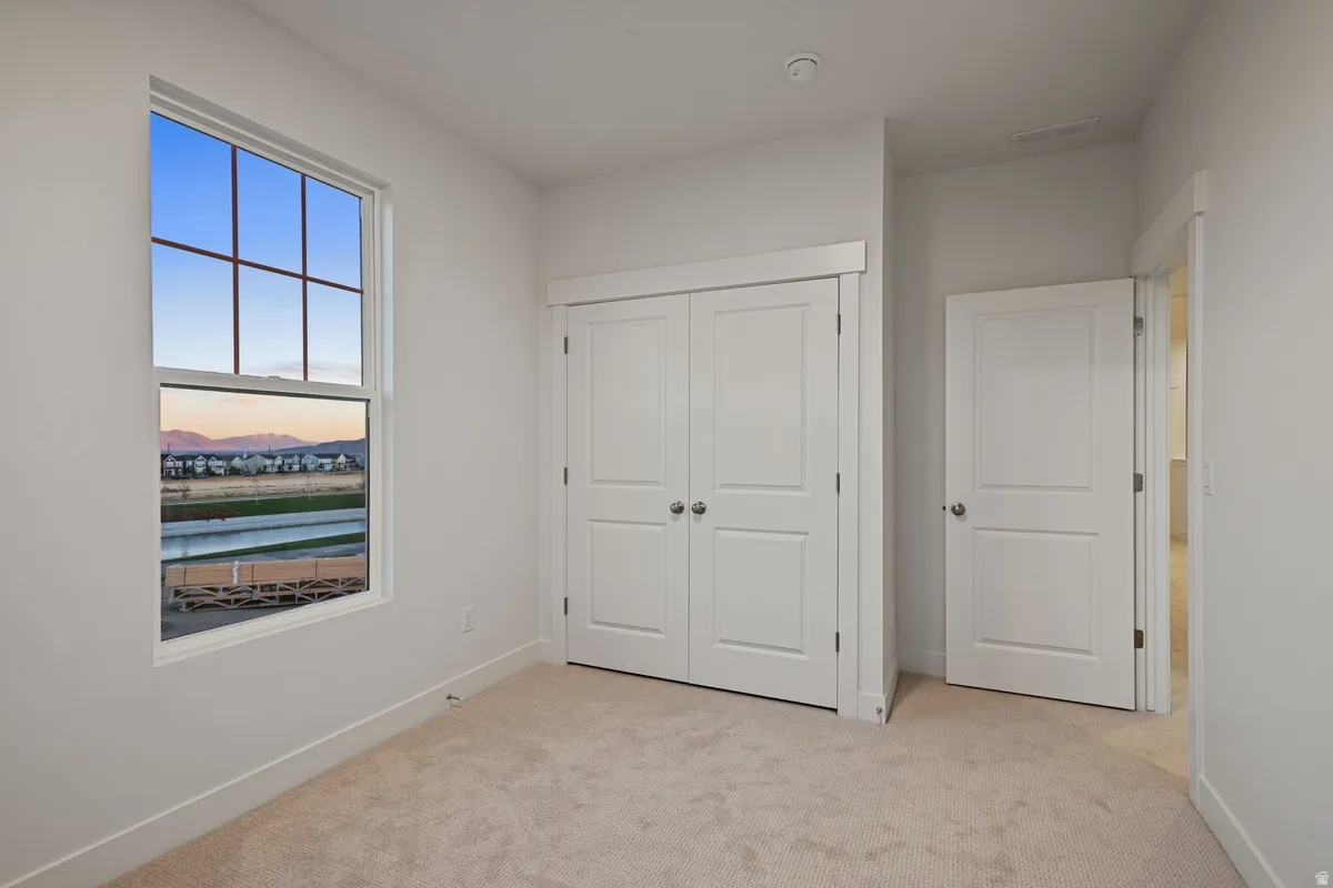 Bright room featuring a large window with grid pattern, neutral-toned carpeting, white walls, and a double-door closet with panel detailing and round door handles