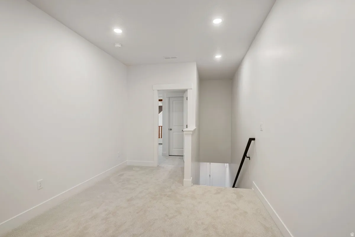 Recessed lighting illuminates a carpeted landing featuring white baseboards, a white door frame, and a dark metal handrail for the descending staircase