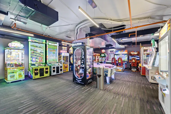 Spacious entertainment area featuring patterned carpet flooring, exposed ductwork, fluorescent lighting, and a variety of arcade-style game machines