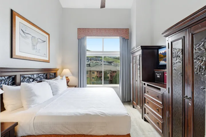 Room featuring wood-finish flooring, a window with drapery, a ceiling fan, a dark wood headboard with carved details, and a dark wood armoire system with carved panels and integrated storage