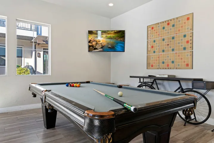 Recreation room featuring a classic billiards table with a light grey felt surface, wood-finish flooring, a wall-mounted flat-screen television, and multiple large windows for natural illumination