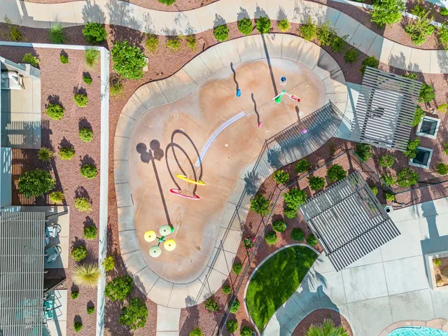 Splash pad with multiple water features, surrounded by pathways, two slatted pergolas, and landscaped grounds with mature shrubbery and decorative rock