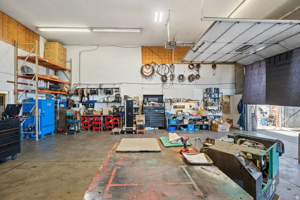 Spacious commercial workshop featuring a concrete floor, high ceilings with fluorescent lighting, industrial shelving, a large overhead door, and ample wall storage