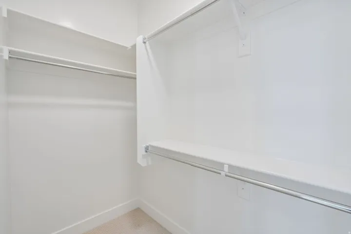 Walk-in closet featuring white walls, white shelving, metal hanging rods, and light-colored carpet flooring