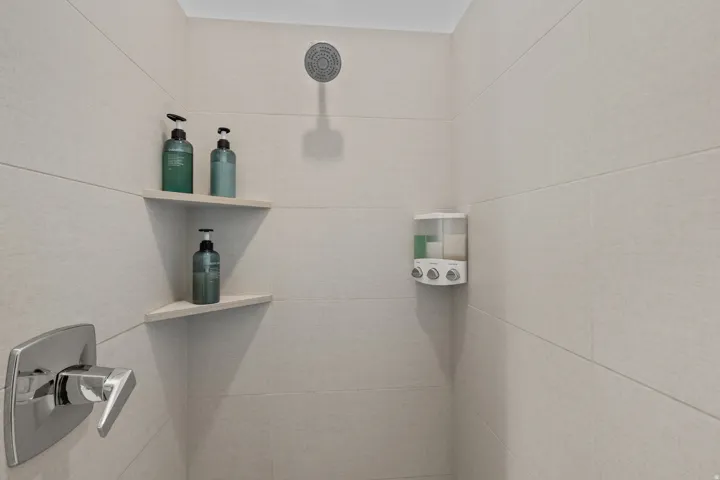 Shower enclosure featuring light-toned rectangular wall tiling, a chrome shower head, a chrome single-handle faucet, and two integrated corner shelves