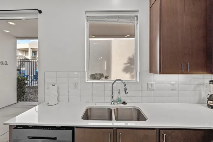 Kitchen featuring a stainless steel double basin sink with a gooseneck faucet, white subway tile backsplash, light-toned countertop, and dark wood-finish cabinetry