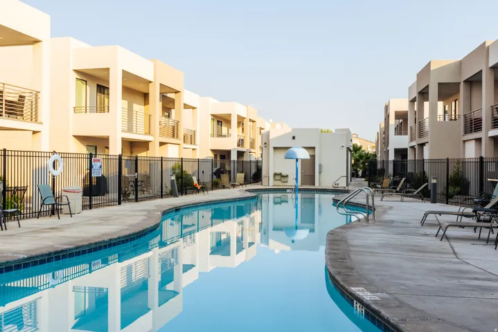 Curved swimming pool with a concrete deck, surrounded by a black metal fence, and modern multi-story buildings featuring private balconies with metal railings