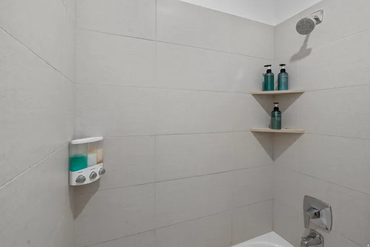 Shower enclosure featuring light-toned tile walls, a chrome shower head, and dual built-in corner shelves
