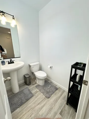Bright powder room featuring a pedestal sink, white toilet, wall-mounted mirror, and wood-finish flooring