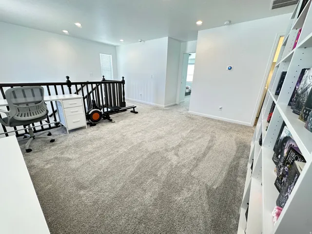 Expansive carpeted loft area featuring a black wood banister, built-in recessed lighting, white baseboards, and a tall built-in shelving unit