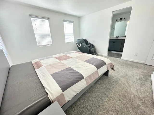 Carpeted room with white walls, two windows featuring horizontal blinds, and an open doorway revealing a bathroom with a vanity and mirror