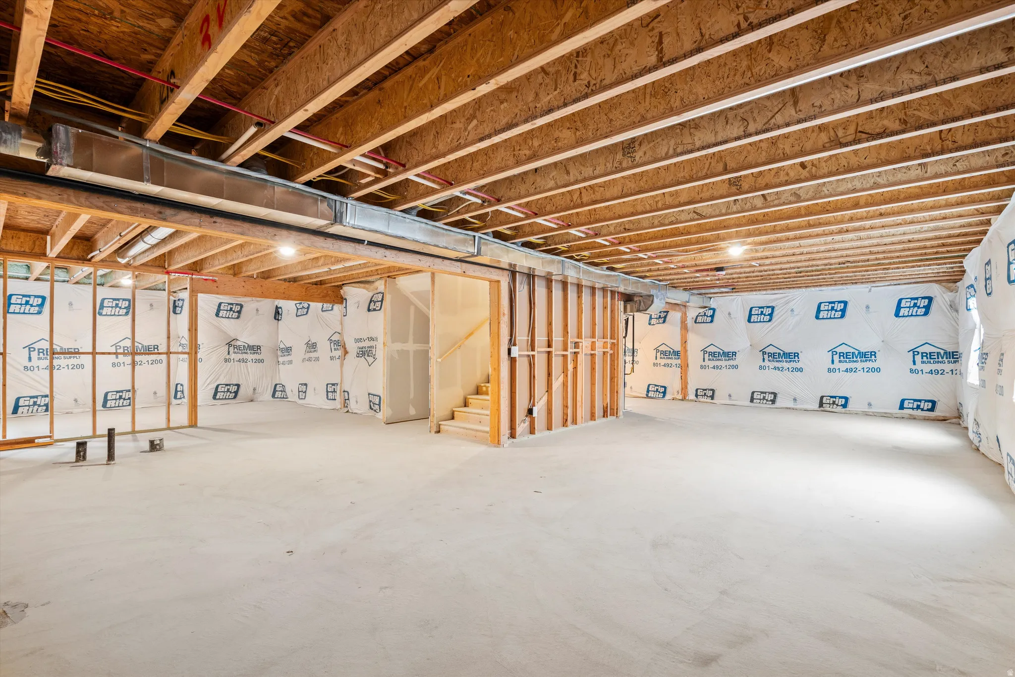 Unfinished lower level featuring exposed wood joists and subflooring, framed walls with insulation wrap, a concrete floor, and a partially finished staircase