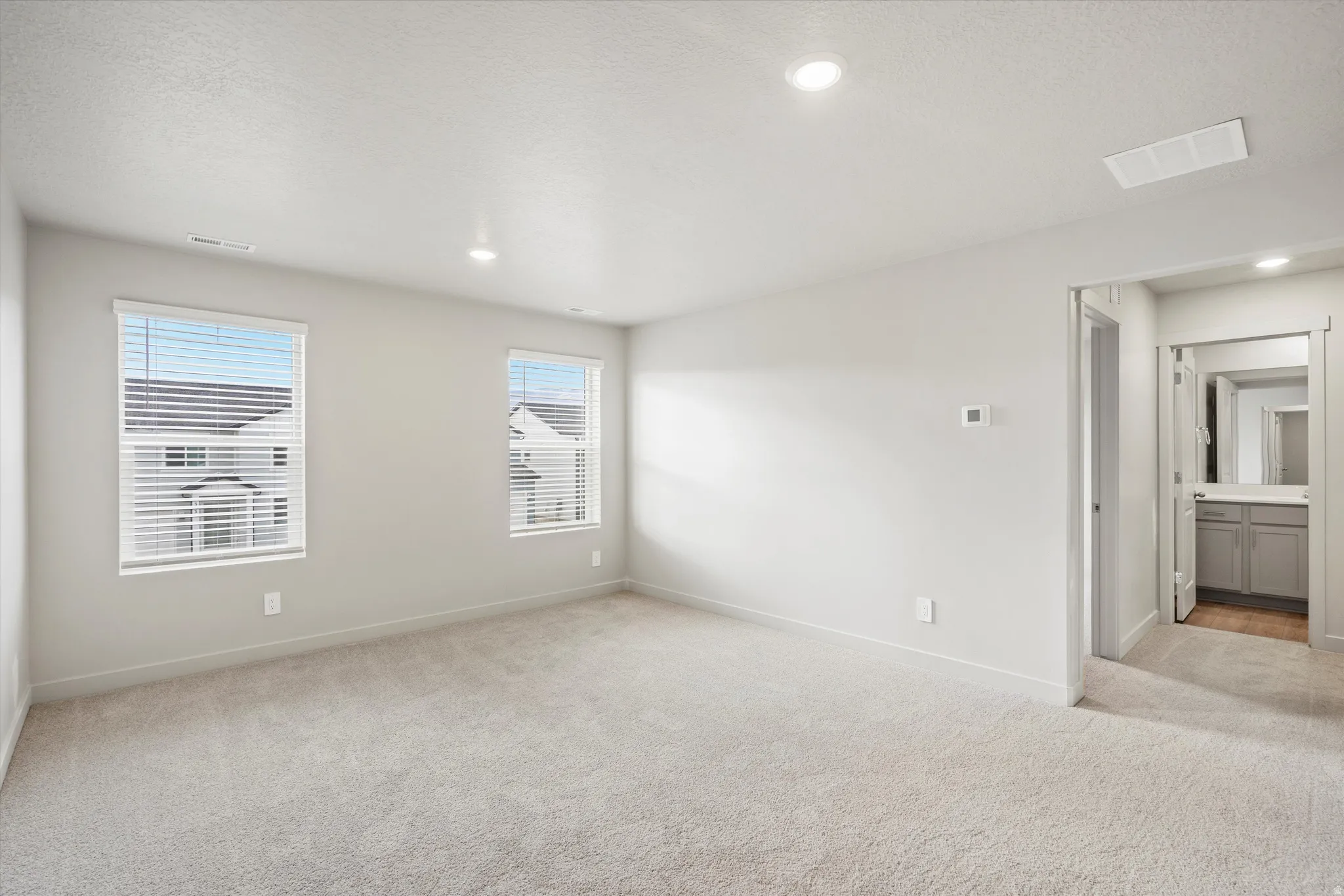 Spacious carpeted room featuring two windows with blinds, recessed lighting, and an entryway to a separate area with a vanity